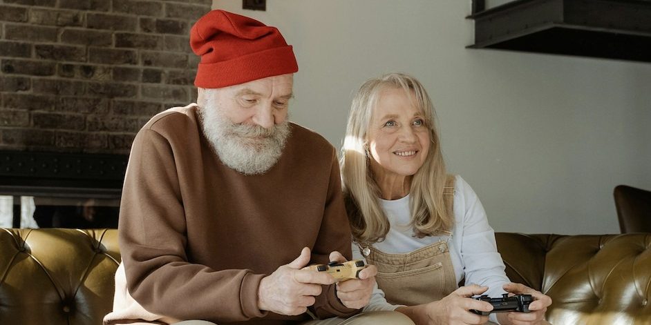 An old man with a red beanie sits on a sofa with an older blonde woman playing video games together.